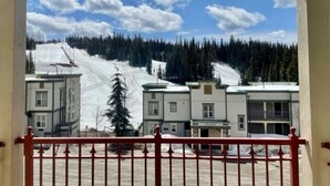 A view from a balcony overlooking a snow-covered slope with buildings in the background. - Creekside condo w/full kitchen, gas fireplace, close to trails & village (Silver Star Mountain)