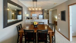 A dining area with a wooden table and chairs, a chandelier, and a kitchen in the background. - Creekside condo w/full kitchen, gas fireplace, close to trails & village (Silver Star Mountain)