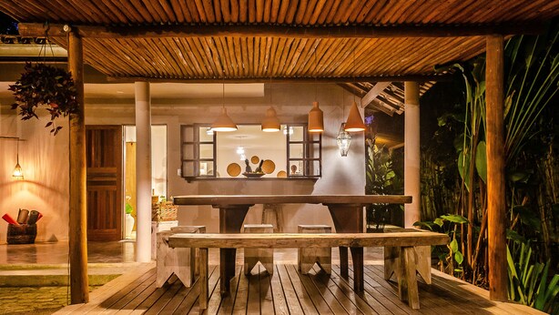Une terrasse extérieure en bois, avec une table et des bancs, entourée d’une végétation luxuriante et surmontée d’une pergola en bois.