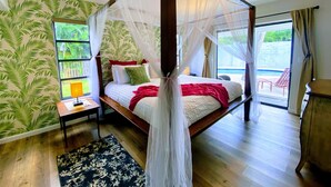 Een bed met hemel, een houten vloer, een wand met een tropisch bladpatroon, een klein nachtkastje en uitzicht op een buitengebied met een zwembad.