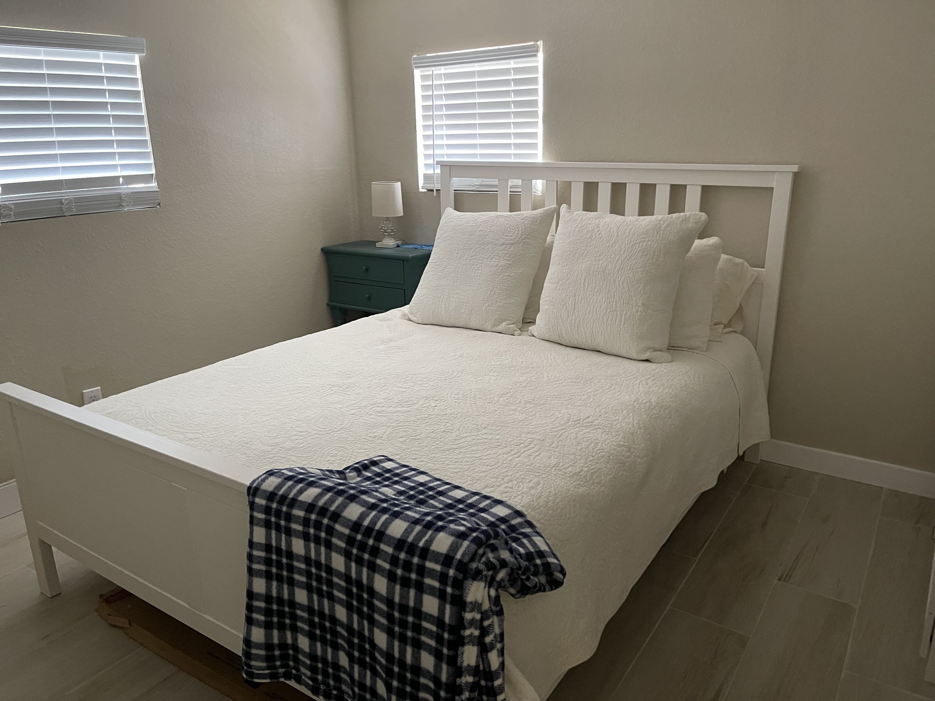 A bedroom with a white bed, two windows with blinds, a nightstand with a lamp, and a plaid blanket on the floor.
