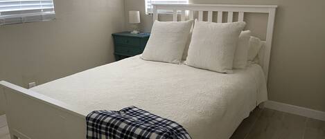 A bedroom with a white bed, two windows with blinds, a nightstand with a lamp, and a plaid blanket on the floor.