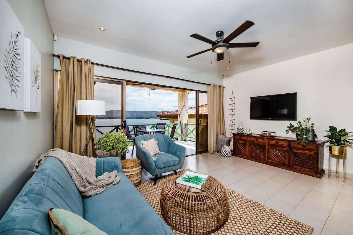 En moderne stue med sofa, lænestol og sofabord. Der er et tv ophængt på væggen, en loftsvifte og en balkon med udsigt.