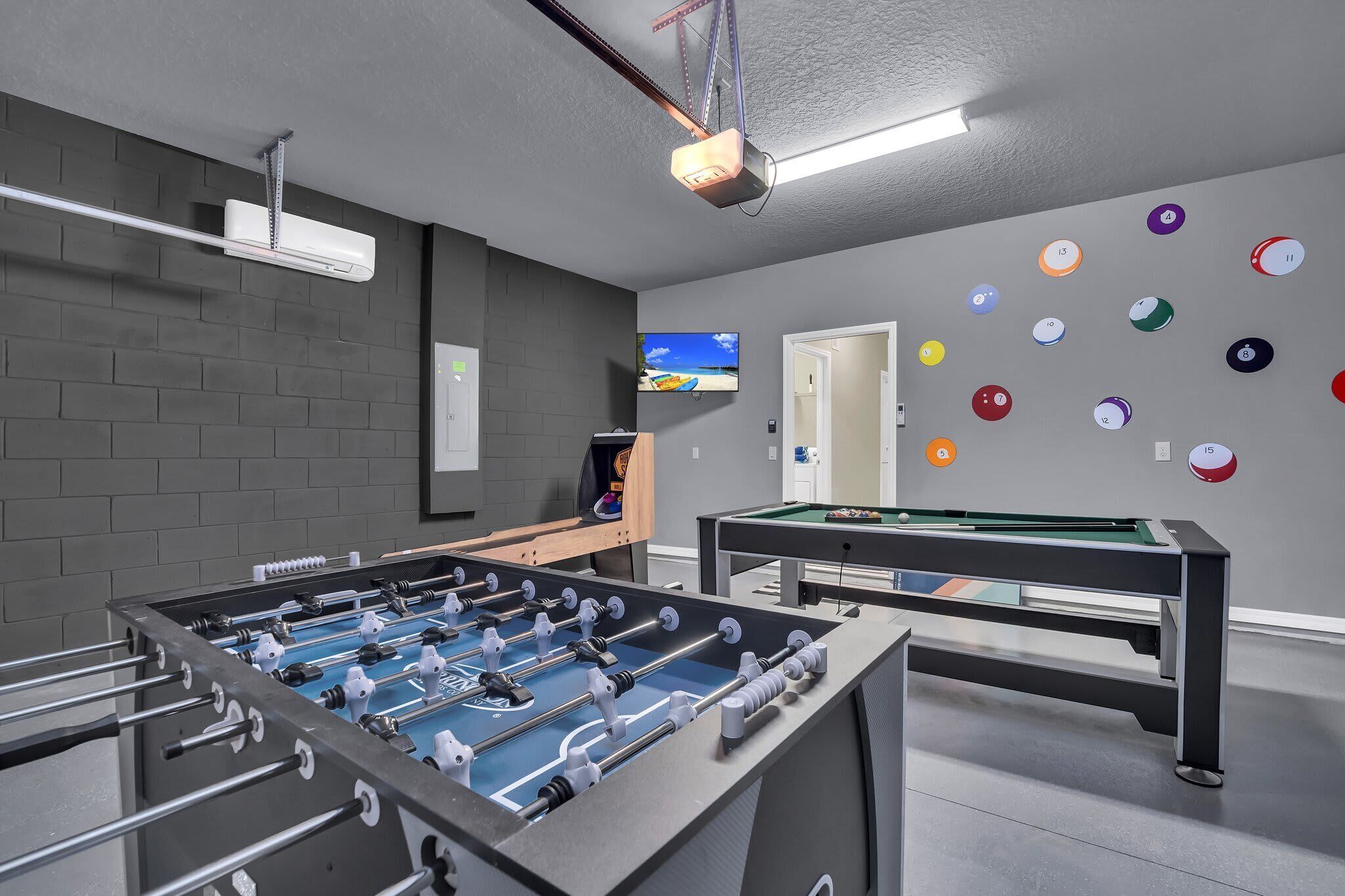 A room with a foosball table, a pool table, and a television mounted on the wall.