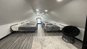 A narrow, dimly lit room with four beds aligned in a row, a small round black chair, and a wooden floor.