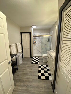 Un baño moderno con ducha, inodoro y lavamanos. Hay una alfombra de cuadros en el suelo y un cesto de ropa cerca de la puerta.