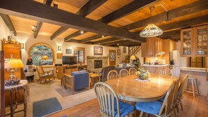 A spacious living area with a large wooden dining table, comfortable seating, a fireplace, and a view of the outdoors through a large window. - Musketeer by Avantstay Family Friendly Home In Tahoe Swiss Village! (Homewood)