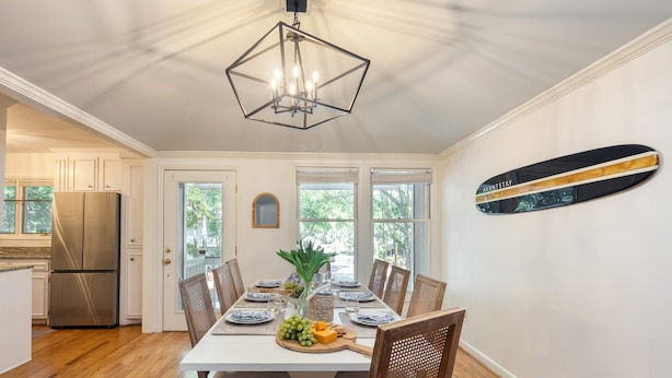 A dining area with a white table set for six, surrounded by wooden chairs. A chandelier hangs from the ceiling, and a skateboard is mounted on the wall. - Crawdad by Avantstay Isle of Palms Home w/ BBQ (Isle of Palms)