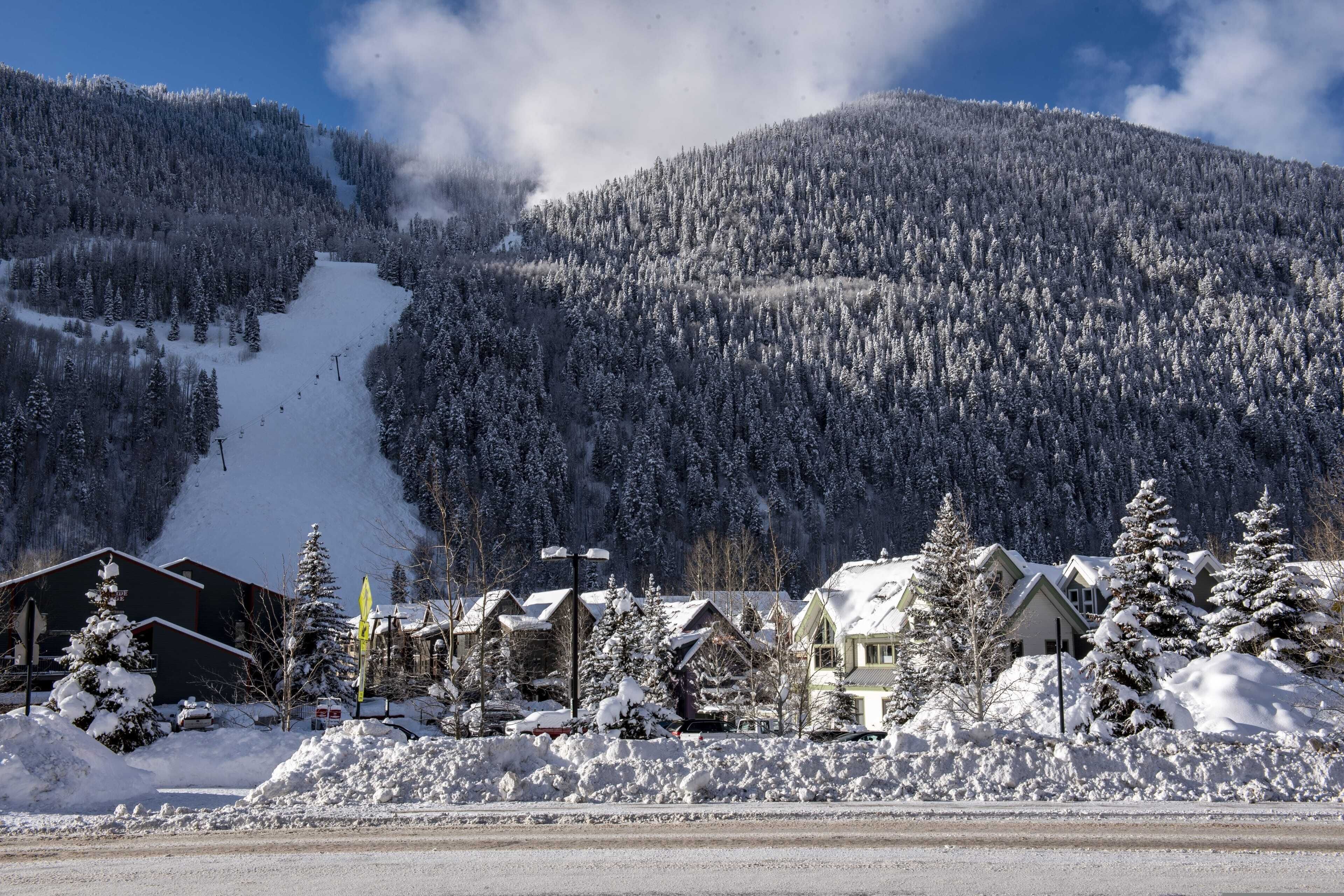 Una montaña nevada con una pista de esquí, una hilera de casas con tejados cubiertos de nieve, y un cielo azul despejado.