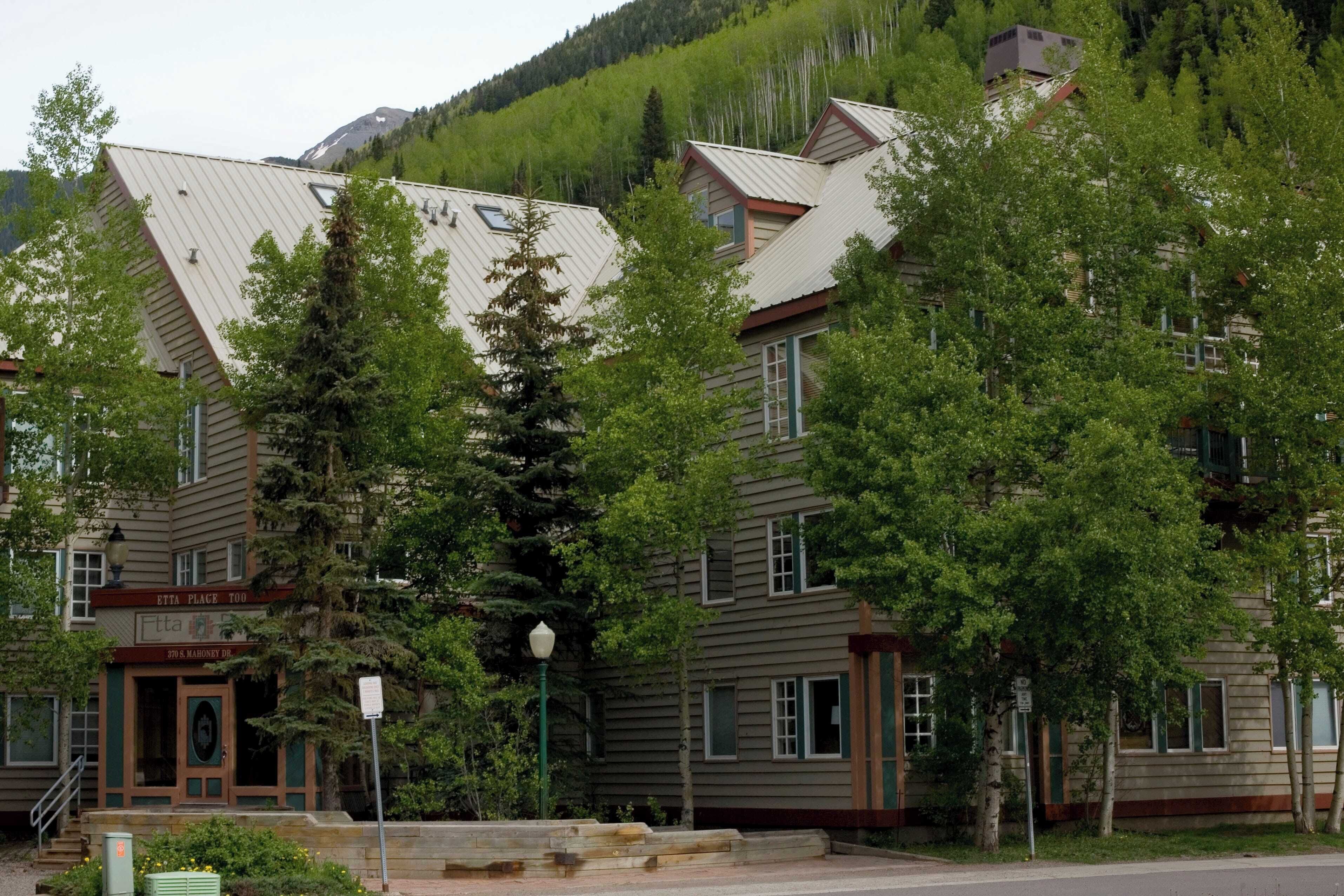 Un immeuble à plusieurs étages, doté d’un toit en métal, entouré d’arbres et se dressant sur un fond de montagnes.