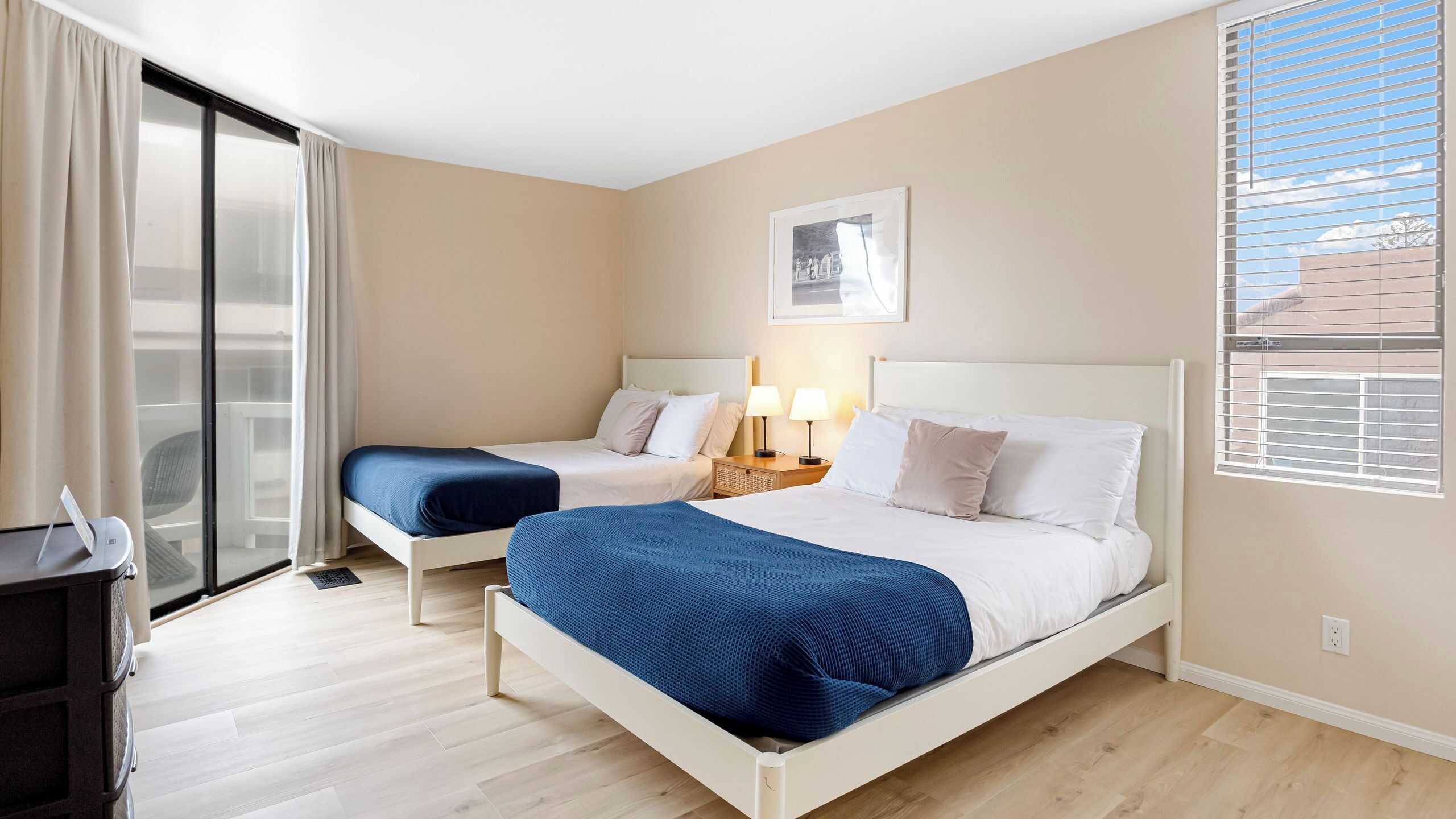 A hotel room with two beds, a desk, a chair, a window with blinds, and a framed picture on the wall.