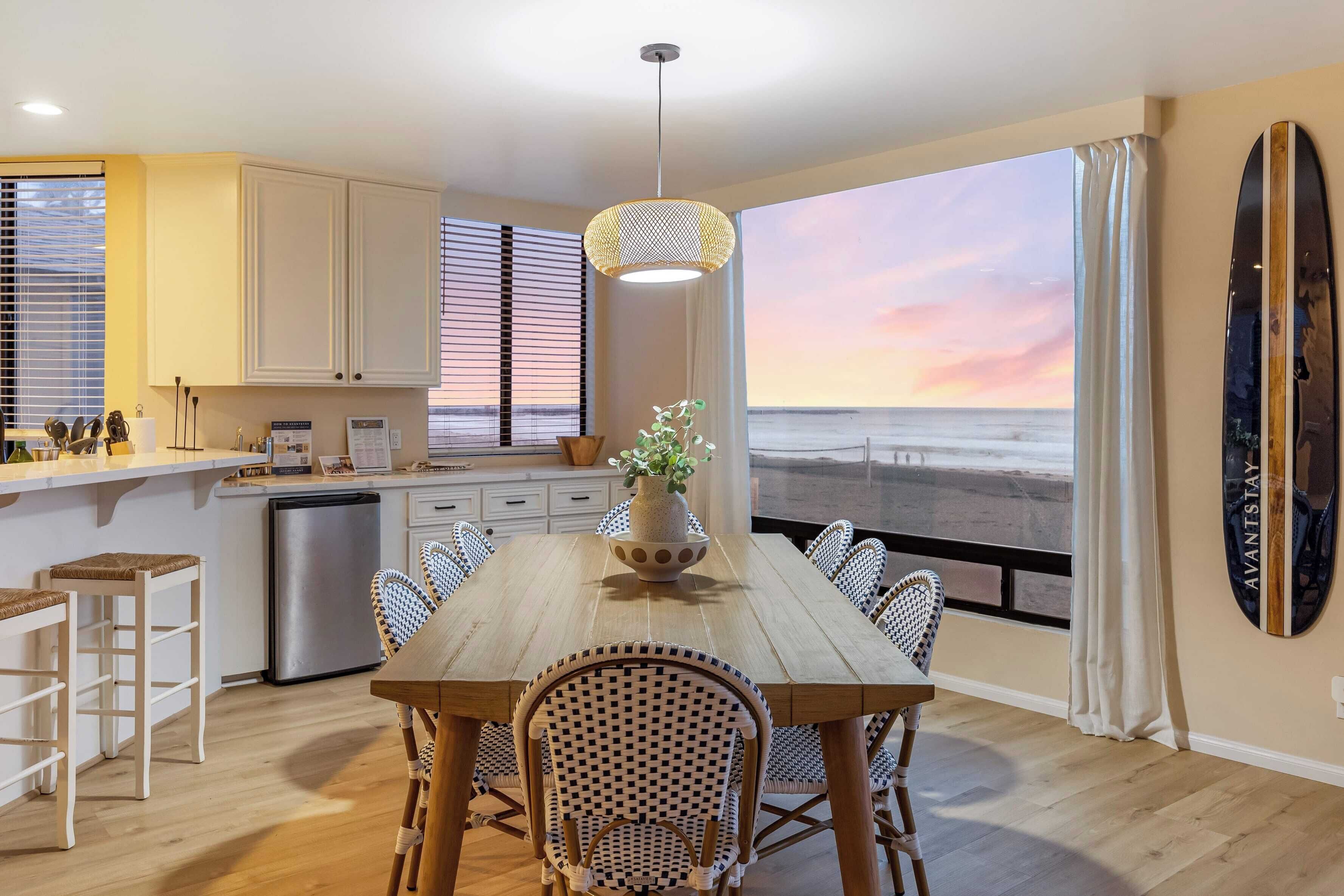 A modern kitchen with white cabinets, a wooden dining table, wicker chairs, and a view of the beach at sunset.
