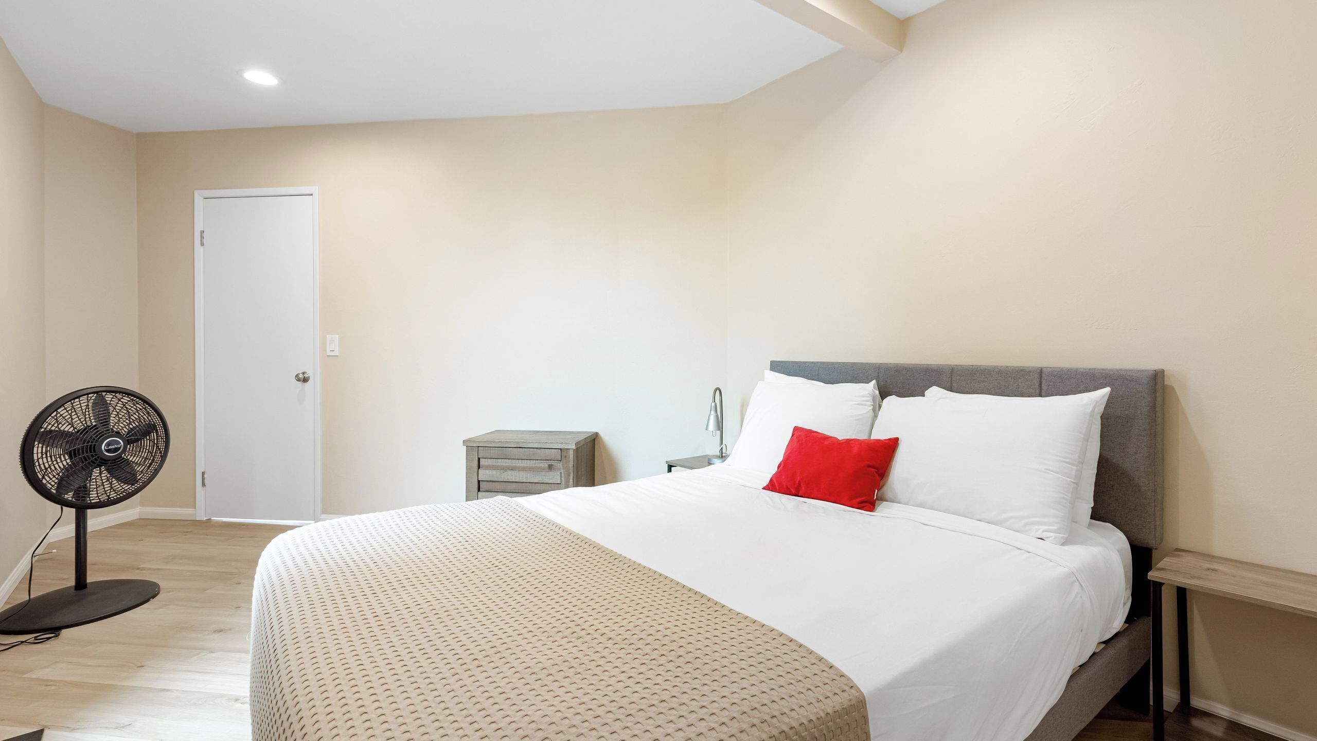 Ein Schlafzimmer mit einem Bett, einem Nachttisch, einer Lampe, einem Ventilator und einer Tür.