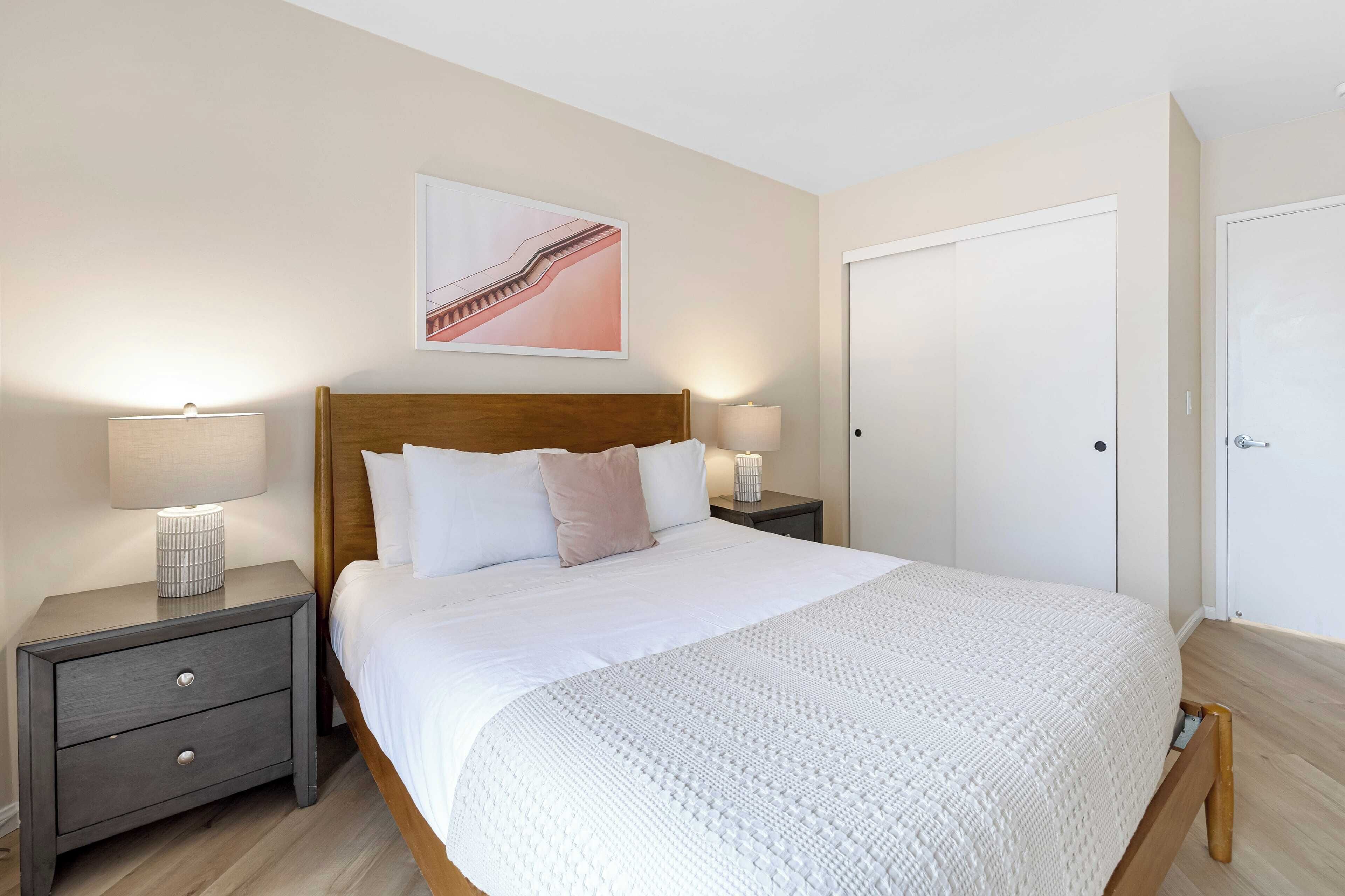 Una cama bien hecha con sábanas blancas, dos mesitas de noche con lámparas, un cabecero de madera y una puerta a la derecha.