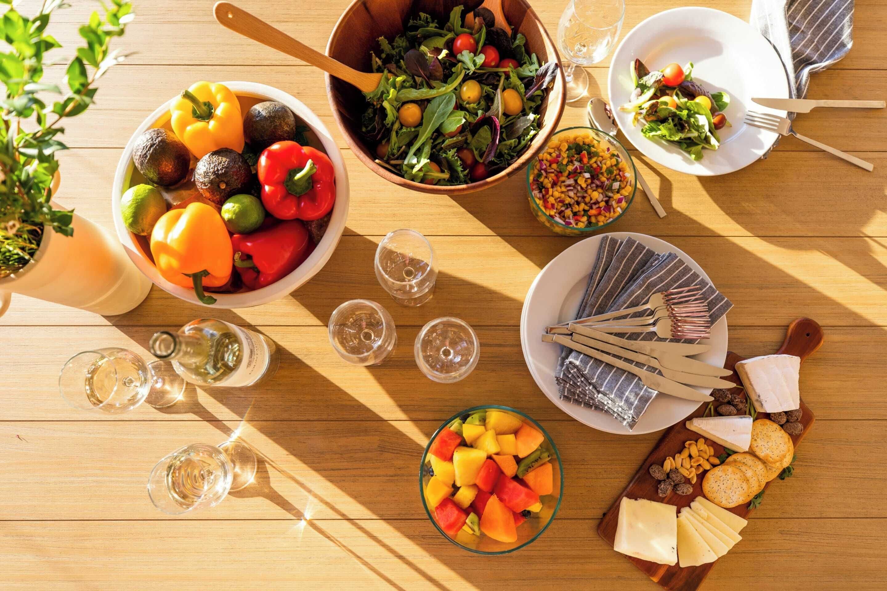 Een houten tafel met diverse etenswaren, waaronder een saladeschaal, een fruitschaal en een bord met kaas.