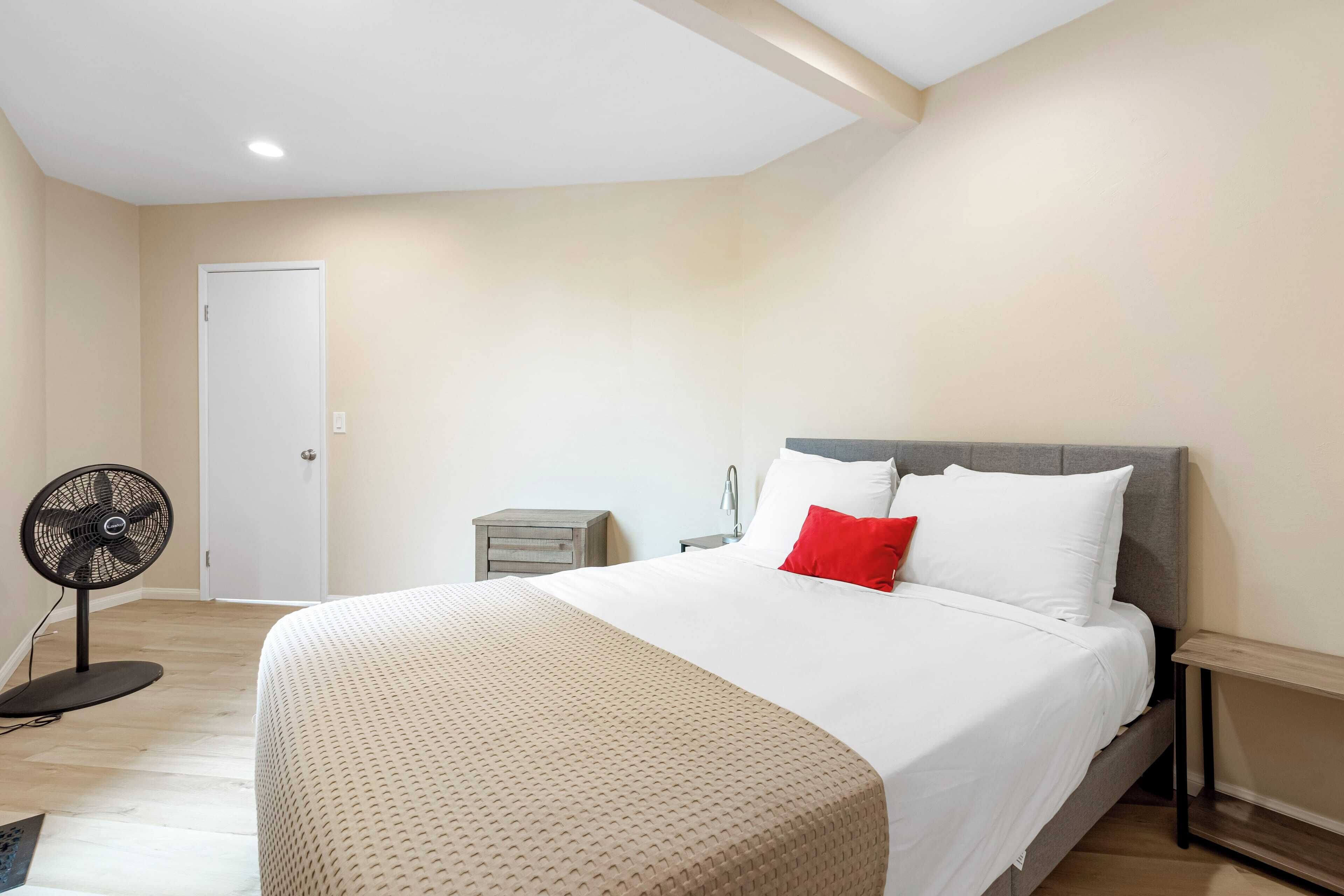 A bedroom with a bed, a bedside table, a nightstand, a fan, and a door.