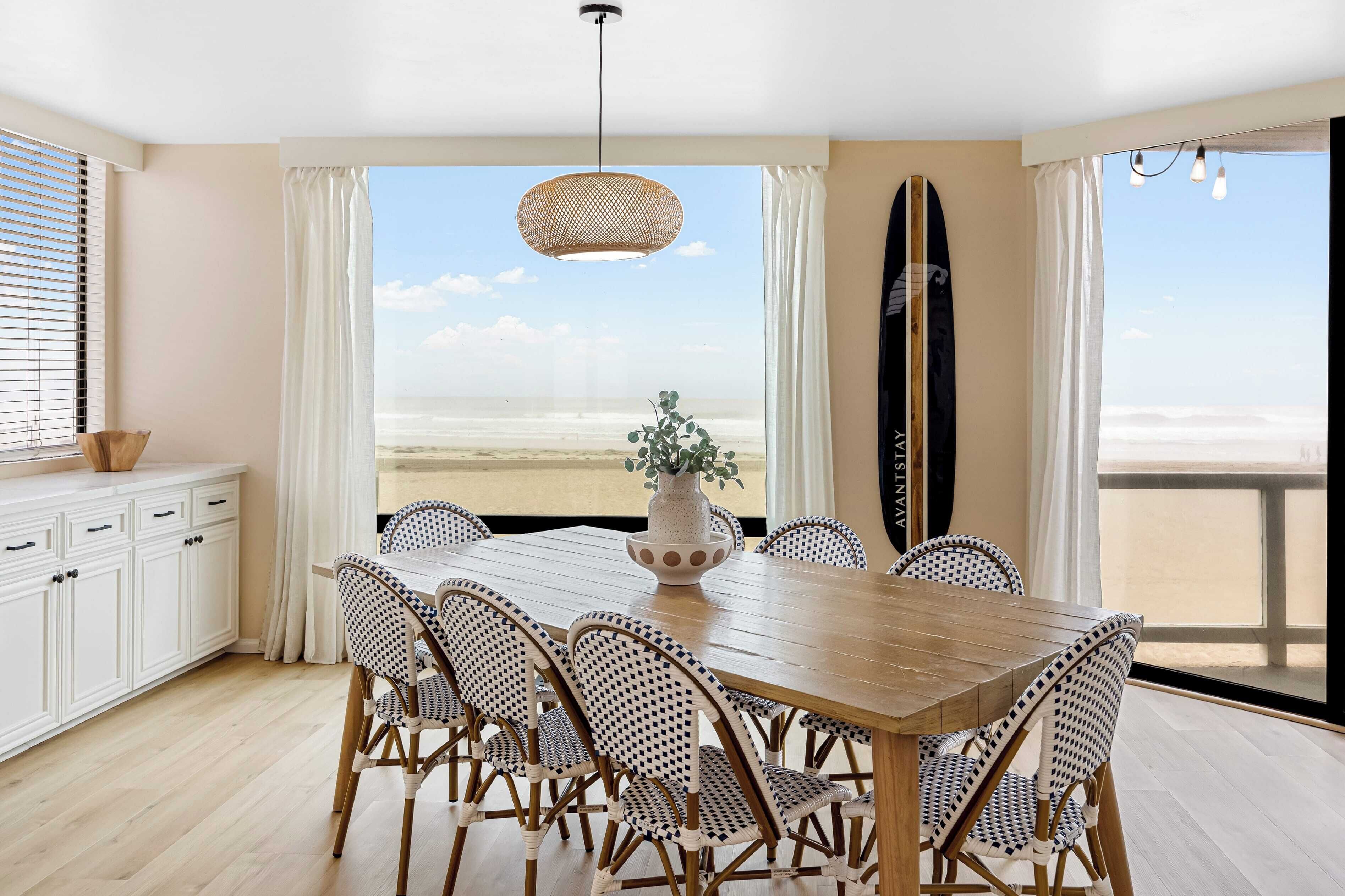 A dining area with a wooden table and wicker chairs, a potted plant, and a surfboard leaning against the wall.