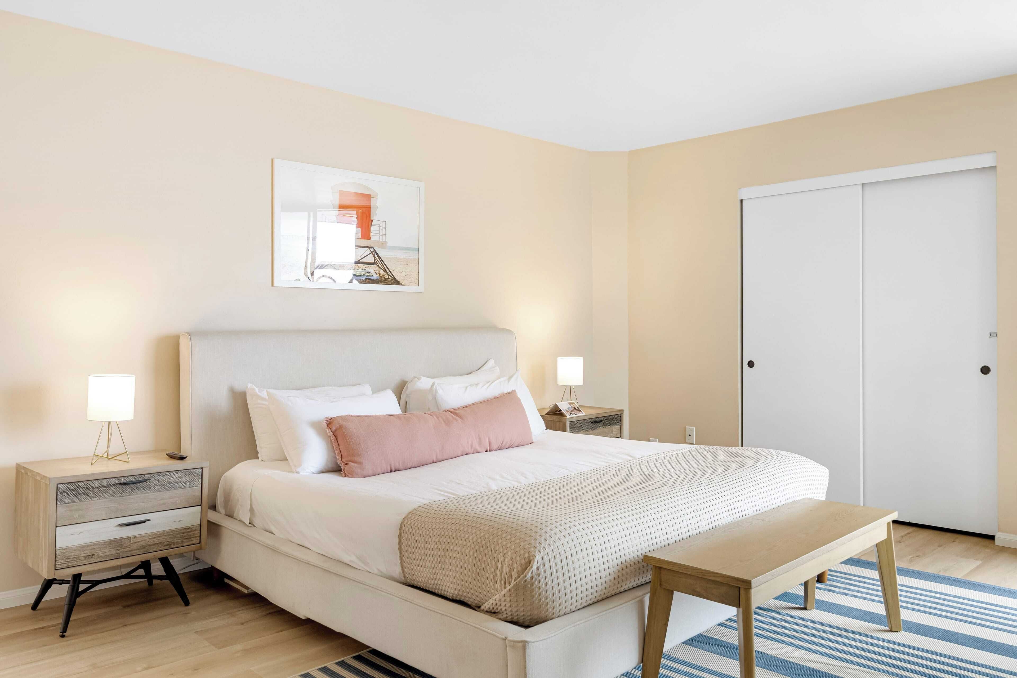 Quarto bem organizado com cama, mesas de cabeceira, um banco e uma porta.