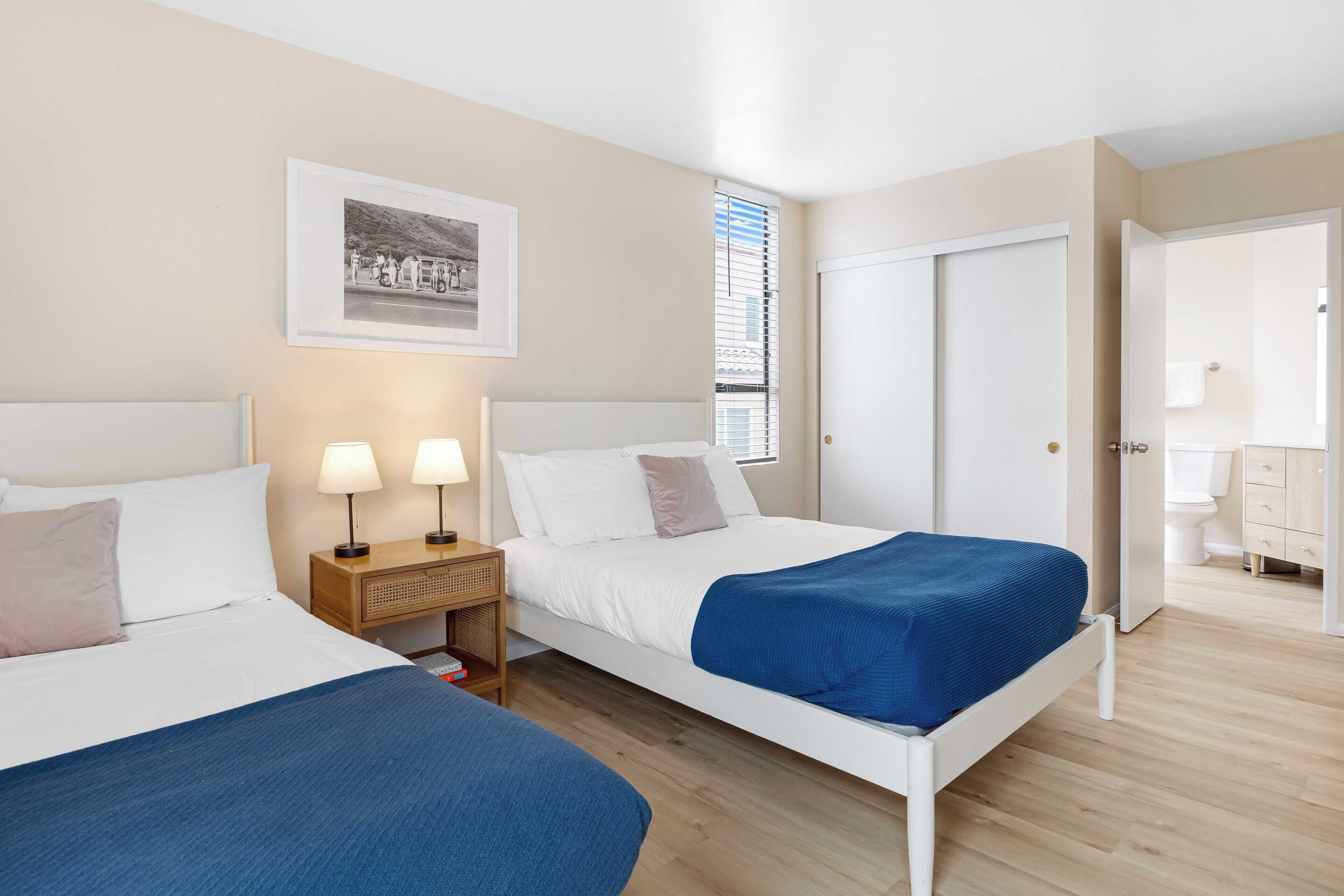 A hotel room with two beds, a bedside table with lamps, a framed picture on the wall, and a view of the bathroom through an open door.