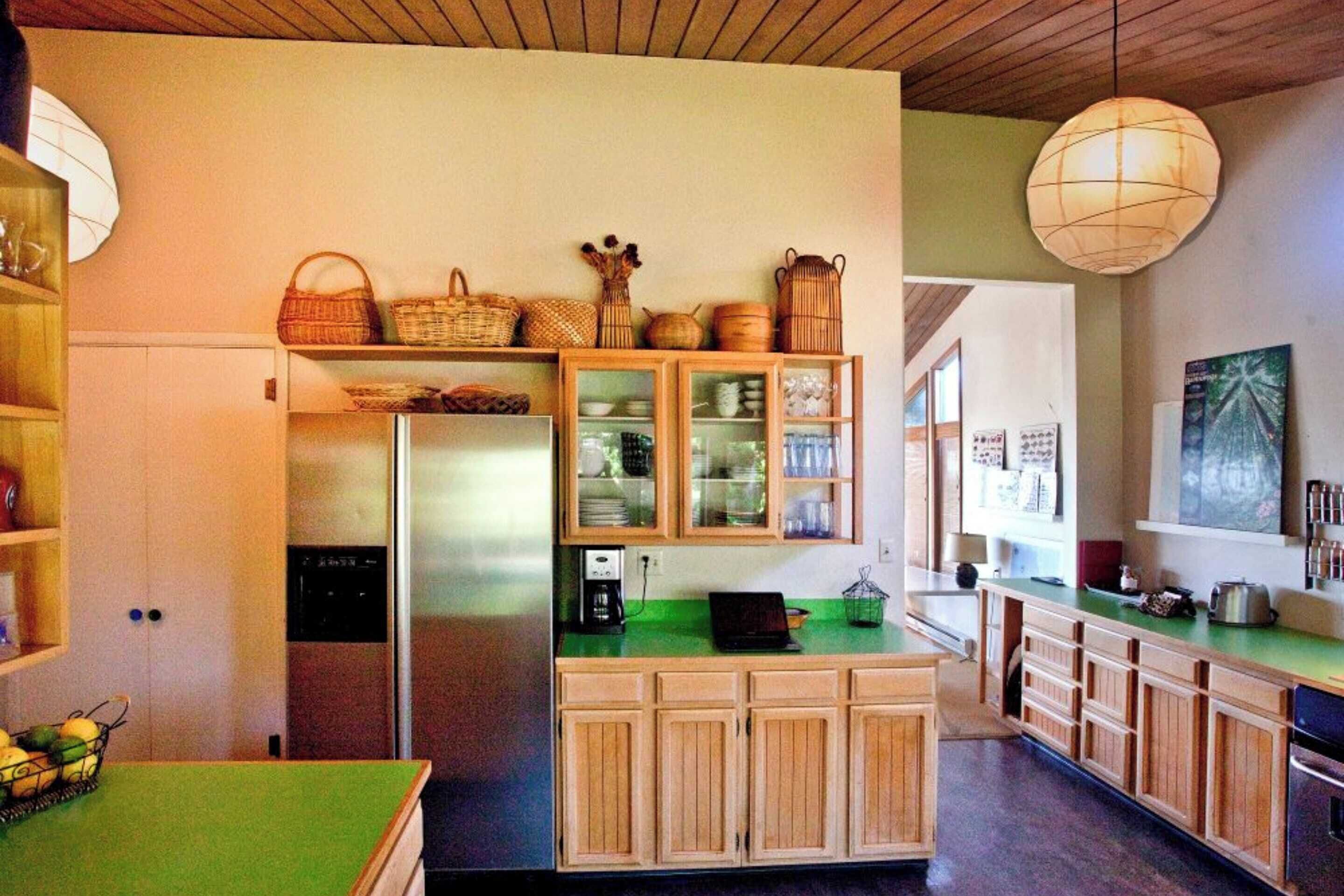 A kitchen with wooden cabinets, a refrigerator, and a green countertop. There are baskets and kitchenware on the shelves.