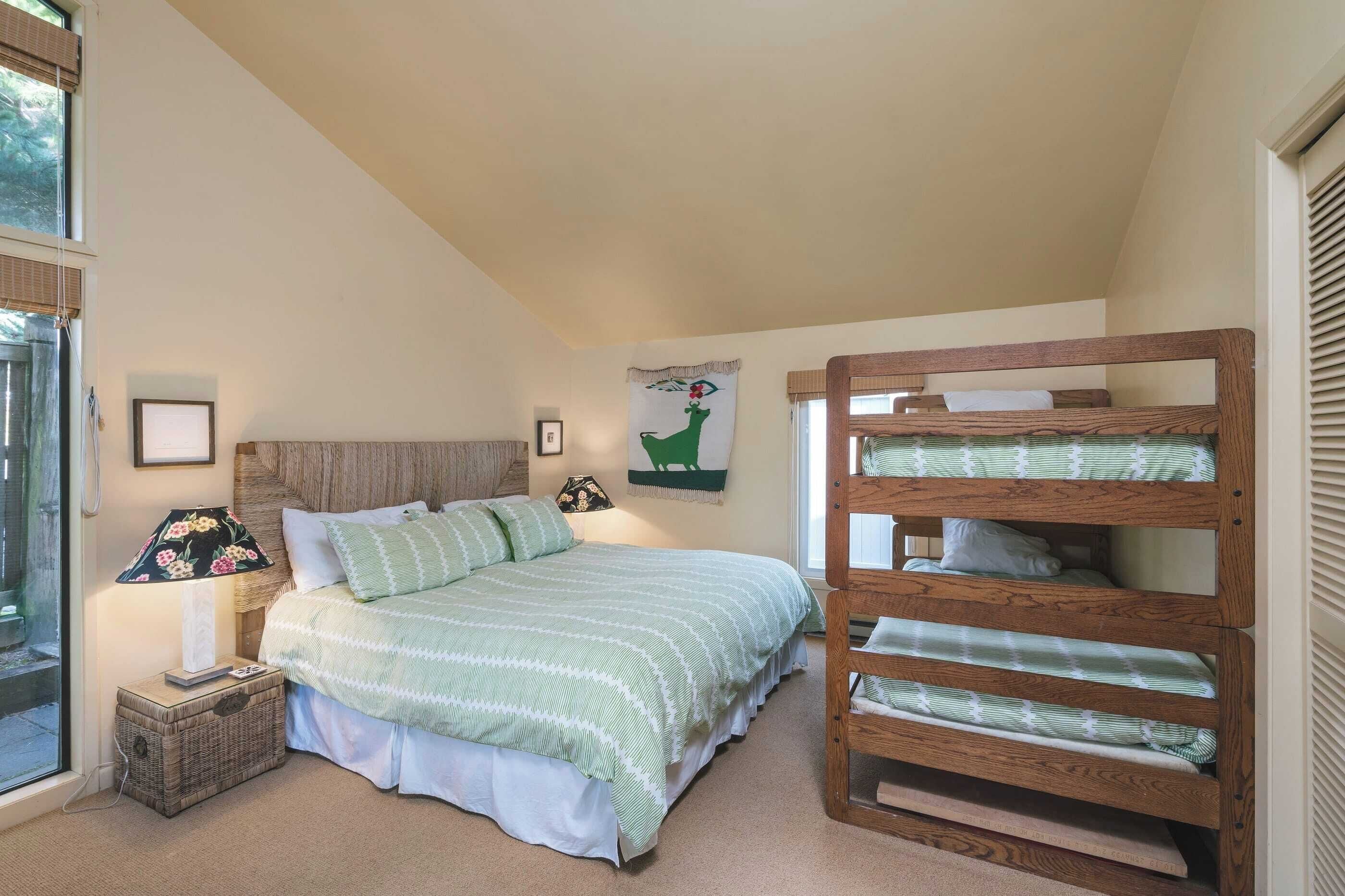 A bedroom with a bed, a wooden bunk bed, a lamp, a framed picture, and a window with blinds.
