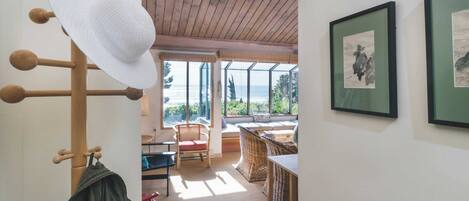 A room with a wooden ceiling, a large window with a view of the sea, a wicker chair, and a table.