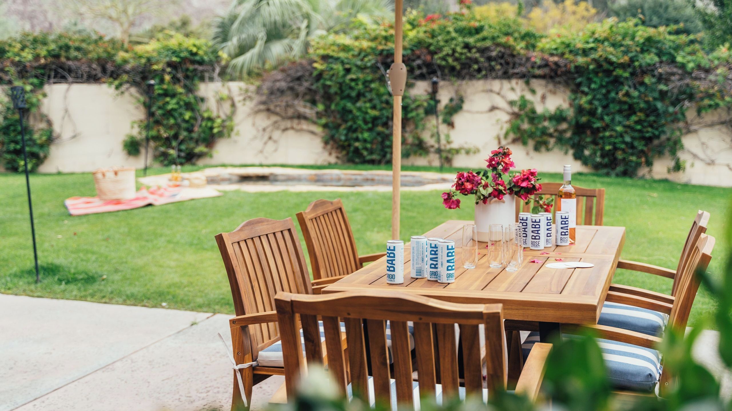 An outdoor wooden table with chairs, a vase with flowers, and cans labeled 'Babe' and 'Grig.'
