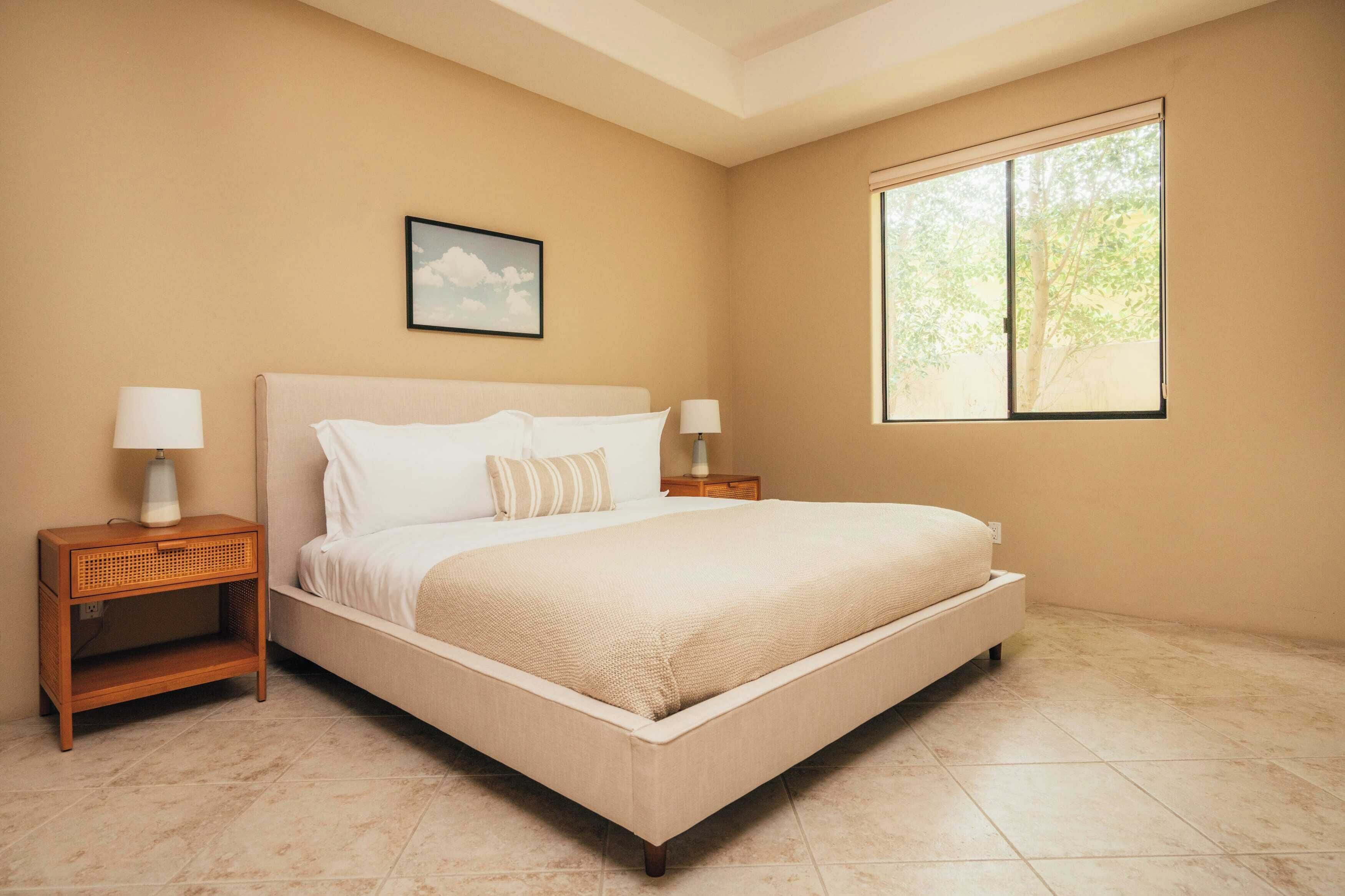 Una camera da letto con un letto grande, due comodini con lampade, una finestra vista alberi e un quadro appeso al muro.