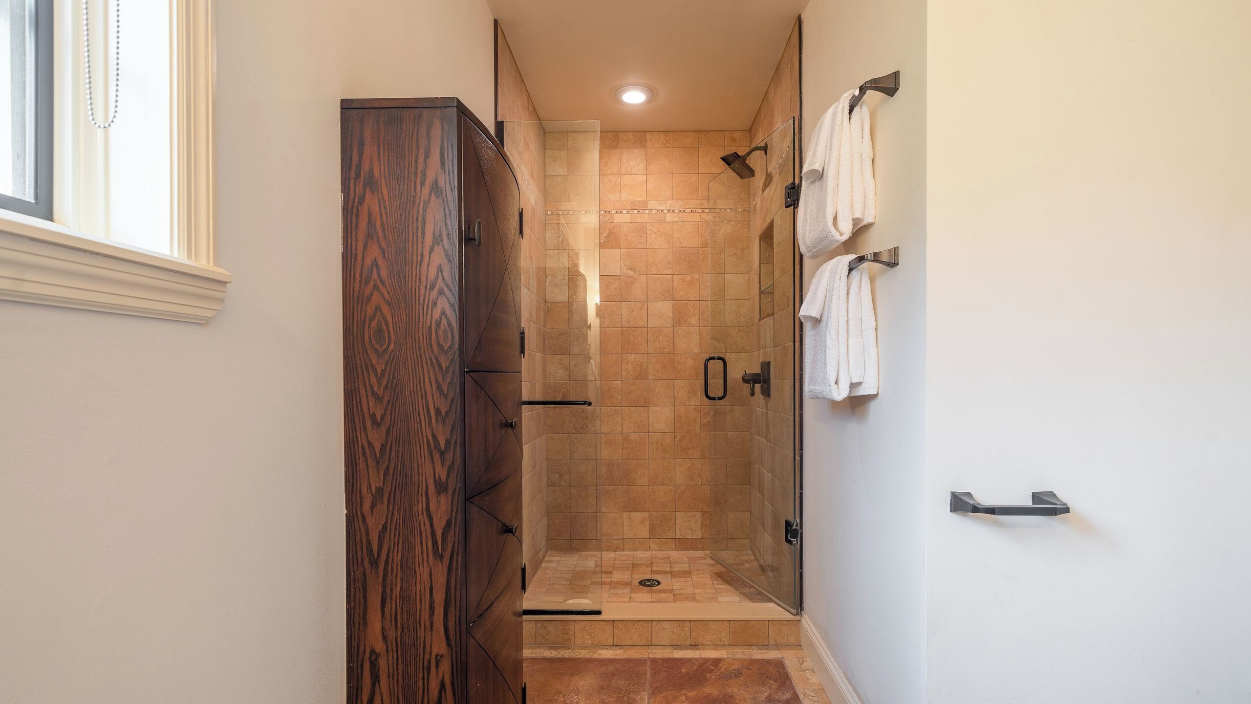 A narrow hallway leading to a shower area with a wooden cabinet and a towel rack.
