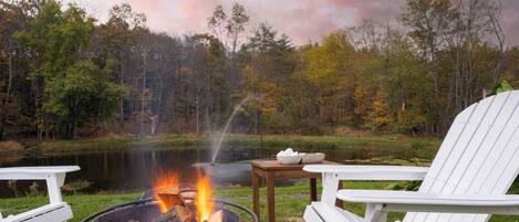A lakeside campsite with a fire pit, white Adirondack chairs, and a small table with bowls.