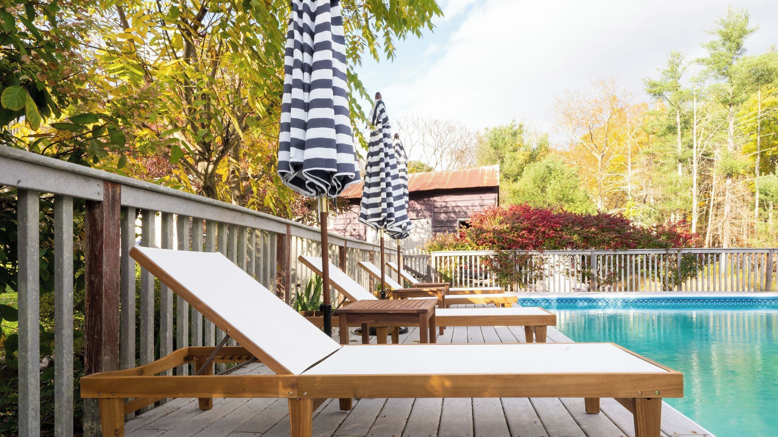 Une terrasse en bois avec des chaises longues et des parasols à rayures, une piscine et une clôture en bois bordée d’arbres et d’arbustes.
