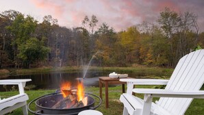 Eine Uferlandschaft mit Feuerstelle, weißen Adirondack-Stühlen und einem Tisch mit einer Schüssel.