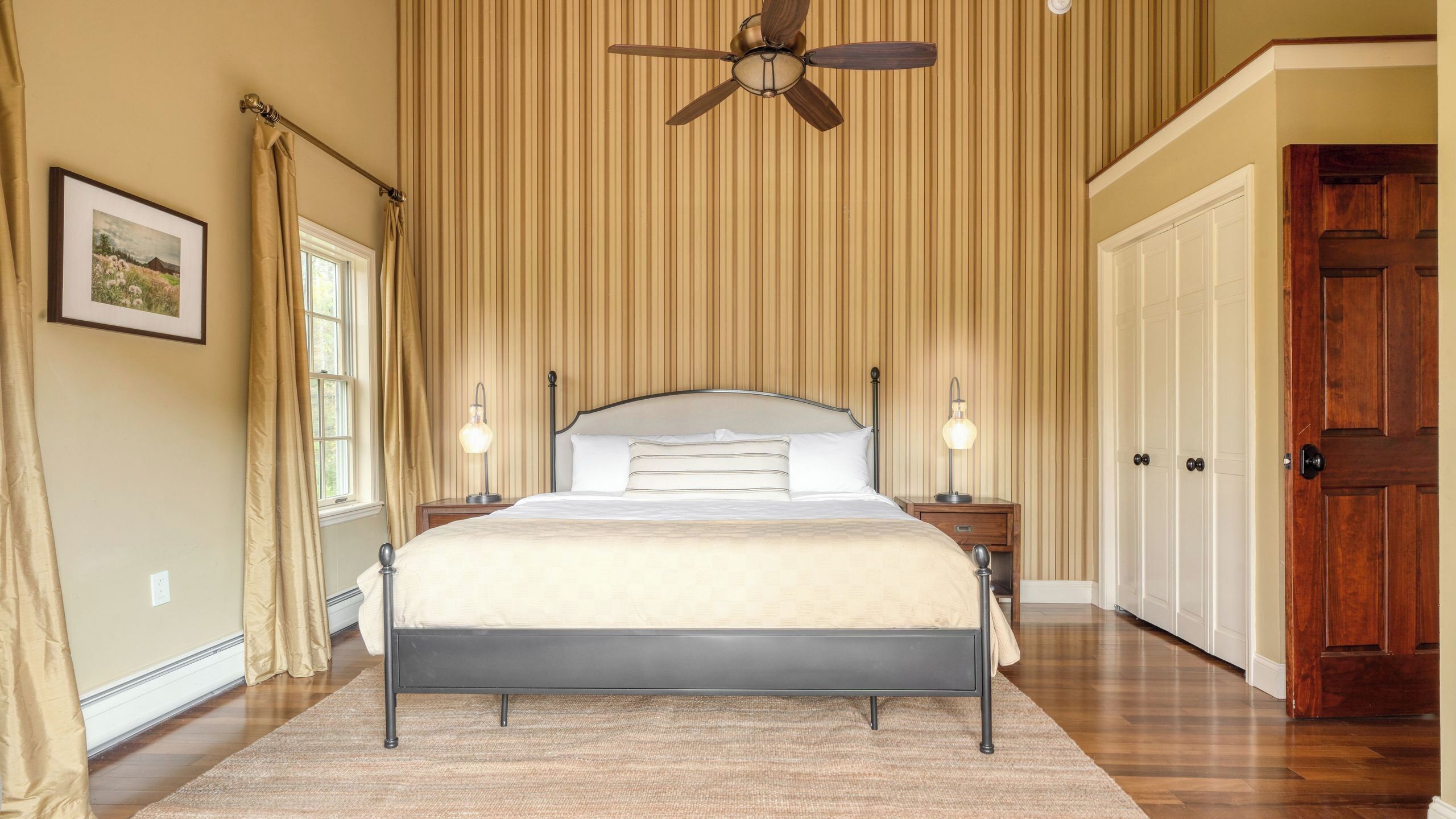 Ein Schlafzimmer mit einem Bett, Deckenventilator, Fenster, Tür und einem gerahmten Bild an der Wand.