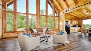 Eine geräumige Holzhütte mit großen Fenstern, einem gemütlichen Wohnbereich mit Sofa und Sesseln, einer Küche mit Holzausstattung und einem Kühlschrank.