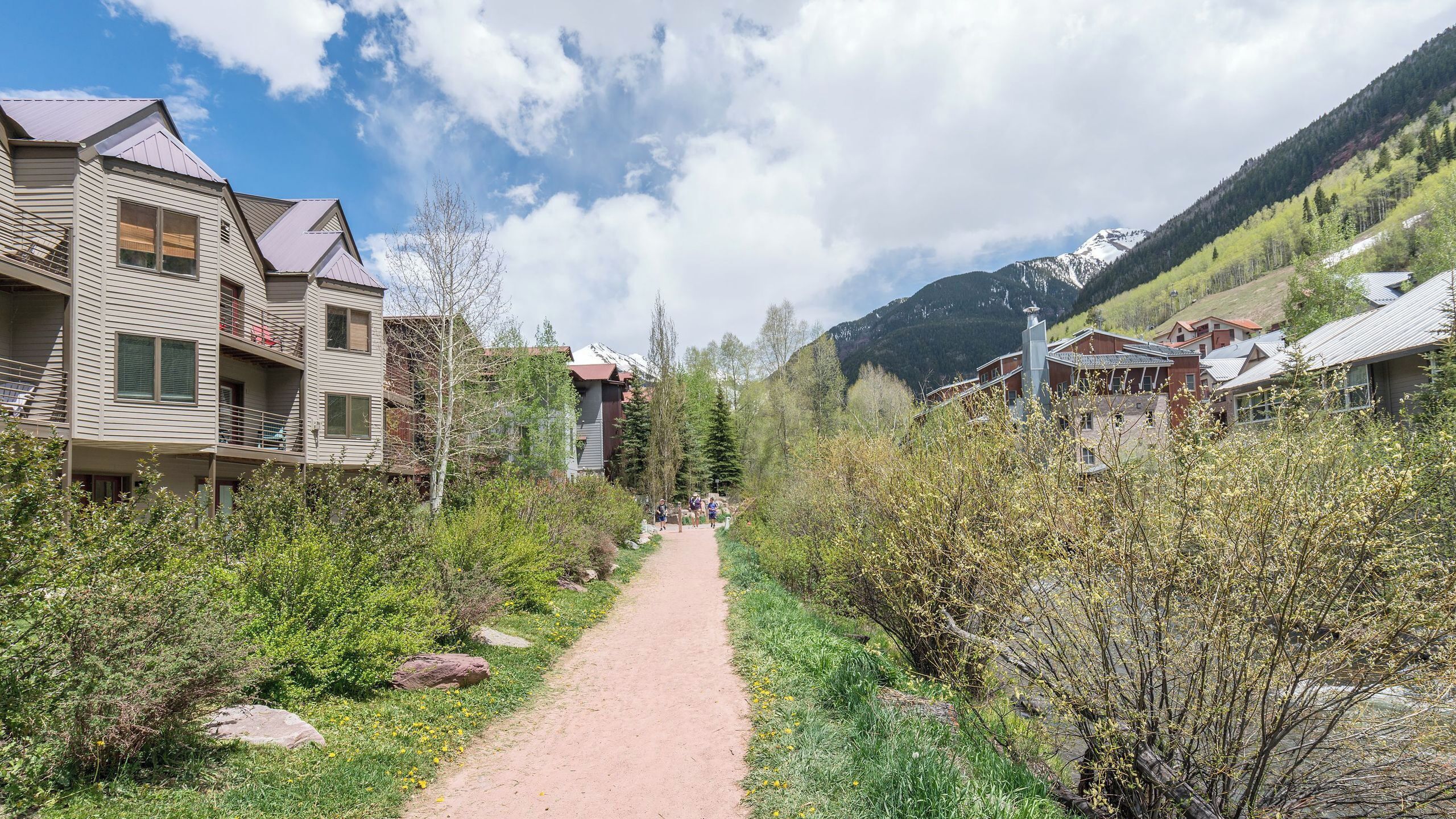 Un sentier qui traverse un quartier résidentiel, bordé de maisons de chaque côté, et entouré de verdure, avec des montagnes en arrière-plan.