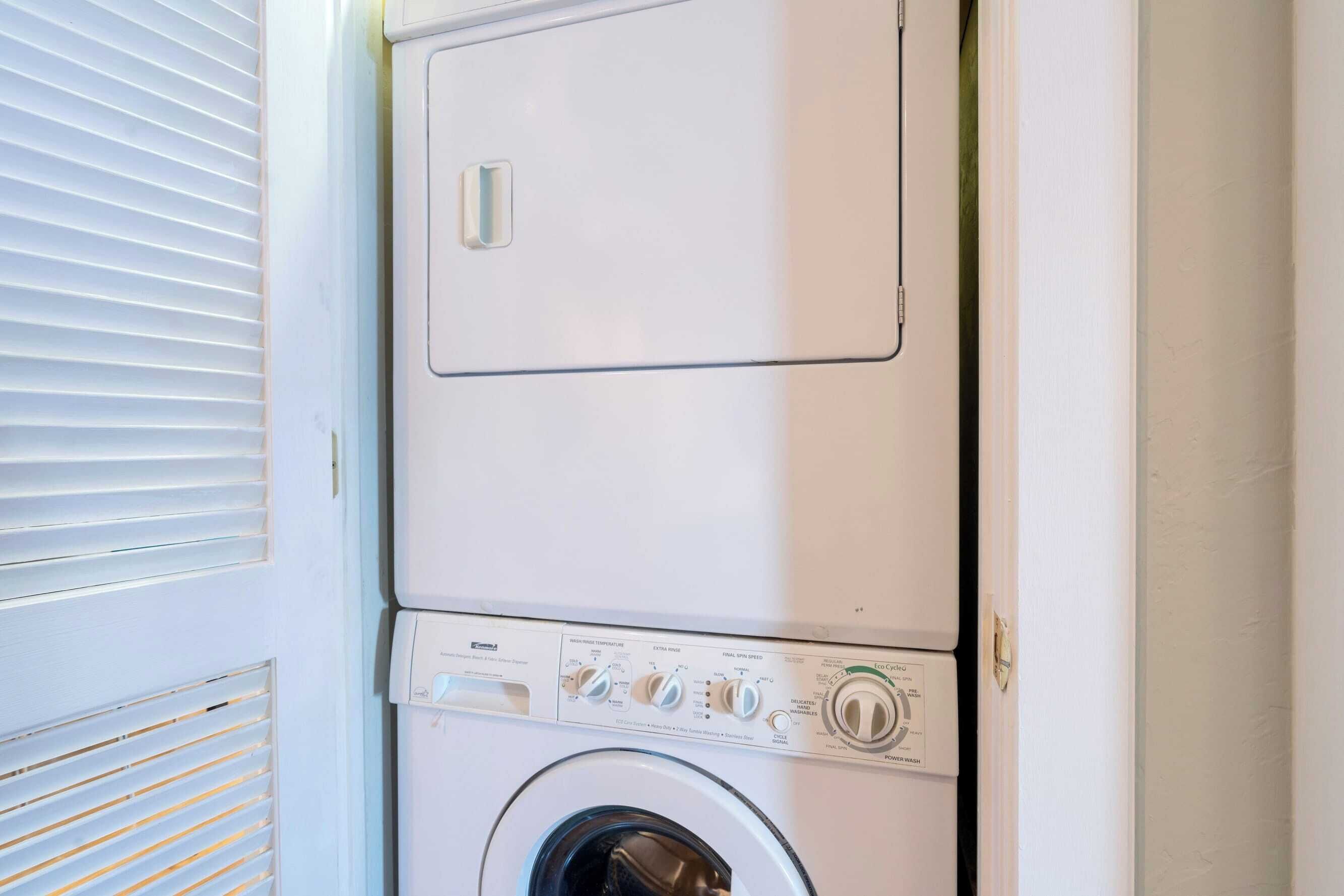 A front-loading washing machine with a control panel, located next to a door with a window with horizontal blinds.
