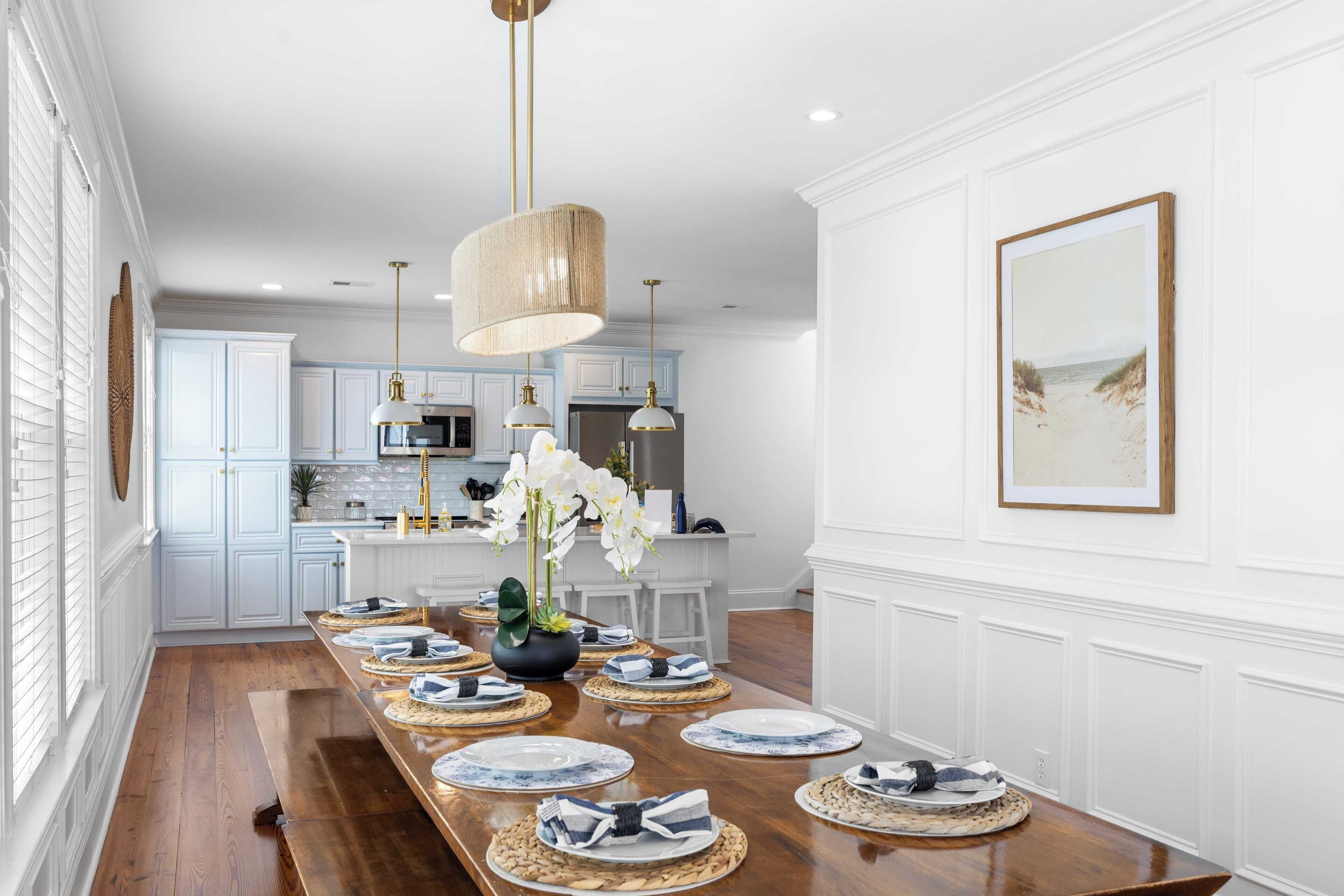 A dining area with a long wooden table set for six, white cabinetry, and pendant lighting.