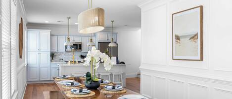 A dining area with a long wooden table set for six, white cabinetry, and pendant lighting.