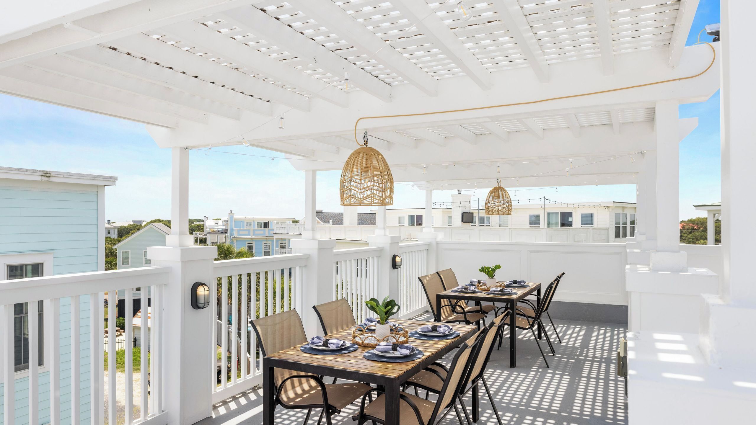 Un patio techado al aire libre con barandilla blanca, una mesa de madera lista para comer y lámparas de mimbre colgantes.