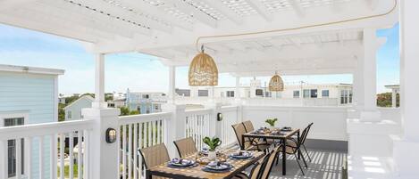Un patio techado al aire libre con barandilla blanca, una mesa de madera lista para comer y lámparas de mimbre colgantes.