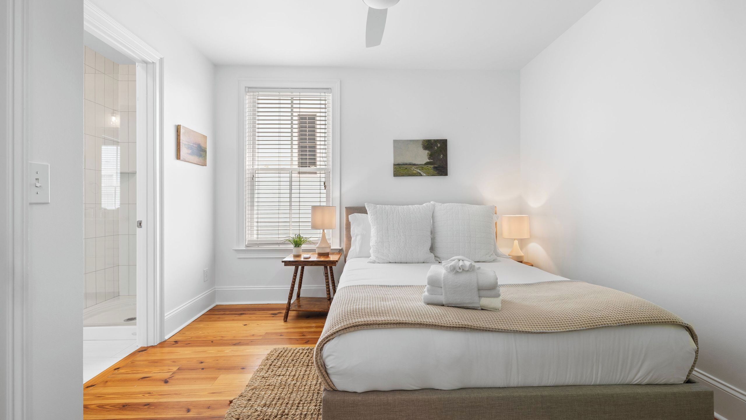 Un dormitorio con cama, lámparas de noche, una mesita con una planta, un cuadro en la pared y una puerta que da a otra habitación.
