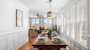 A dining area with a long wooden table set for six, white paneled walls, a ceiling fan, and large windows with blinds. - 1 Block to Beach, Newly Renovated 6bdr w/ Rooftop Views Sand Dune by Avantstay (Isle of Palms)