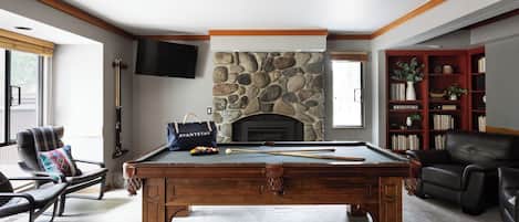 A pool table with a stone fireplace, a bookshelf, and a leather chair.