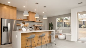A modern kitchen with a central island, bar stools, a stainless steel refrigerator, wooden cabinets, pendant lighting, and a view of the backyard. - Aria by Avantstay Spectacular & Secluded Desert Oasis w/ Pool (Yucca Valley)