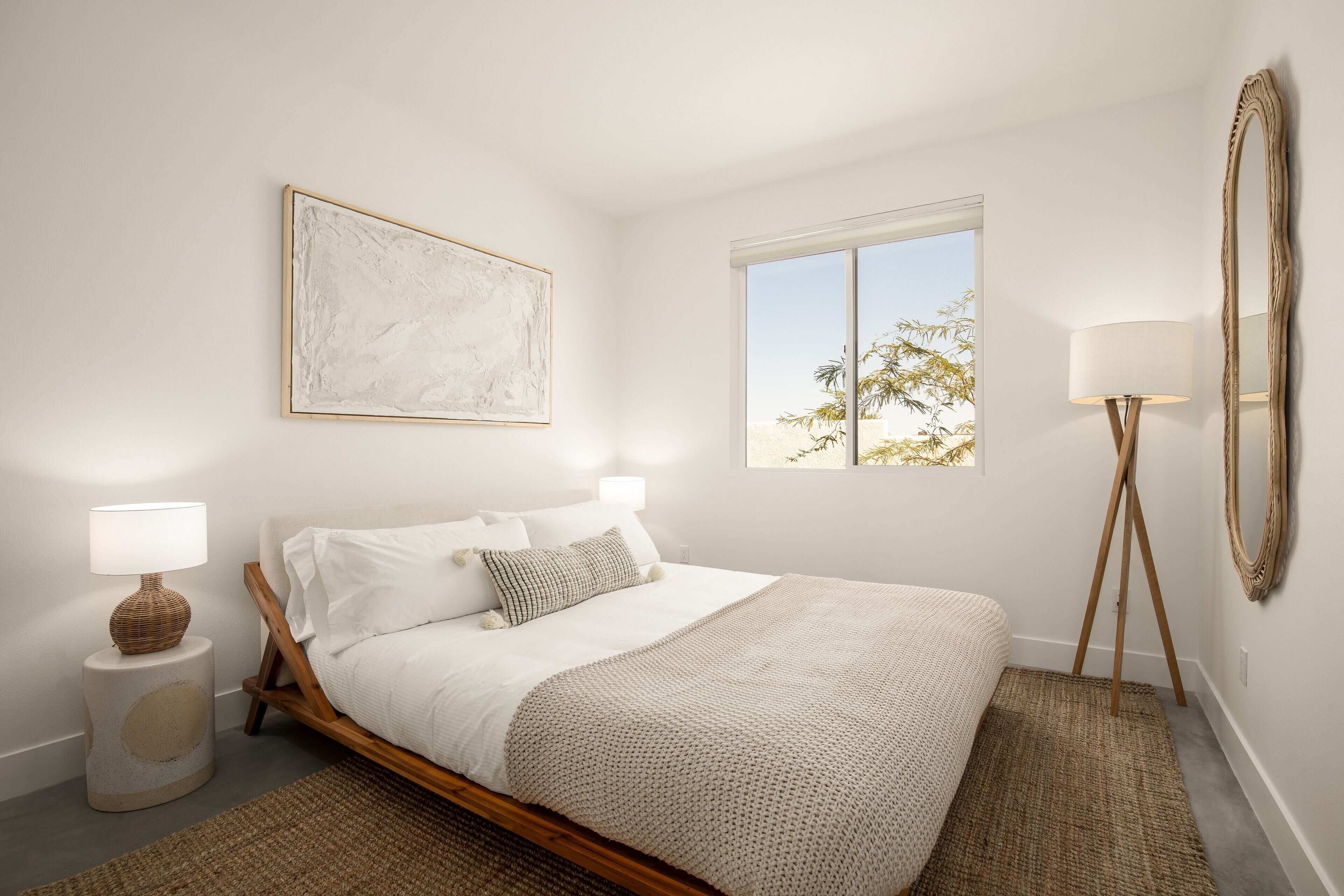 A bedroom with a bed, a bedside lamp, a floor lamp, a mirror, and a window with a view of palm trees.