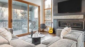 A modern living room with a fireplace, a large TV, and a view of a snowy landscape through large windows.