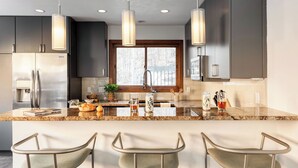 A modern kitchen with a central island, stainless steel appliances, pendant lighting, and a view through a window.