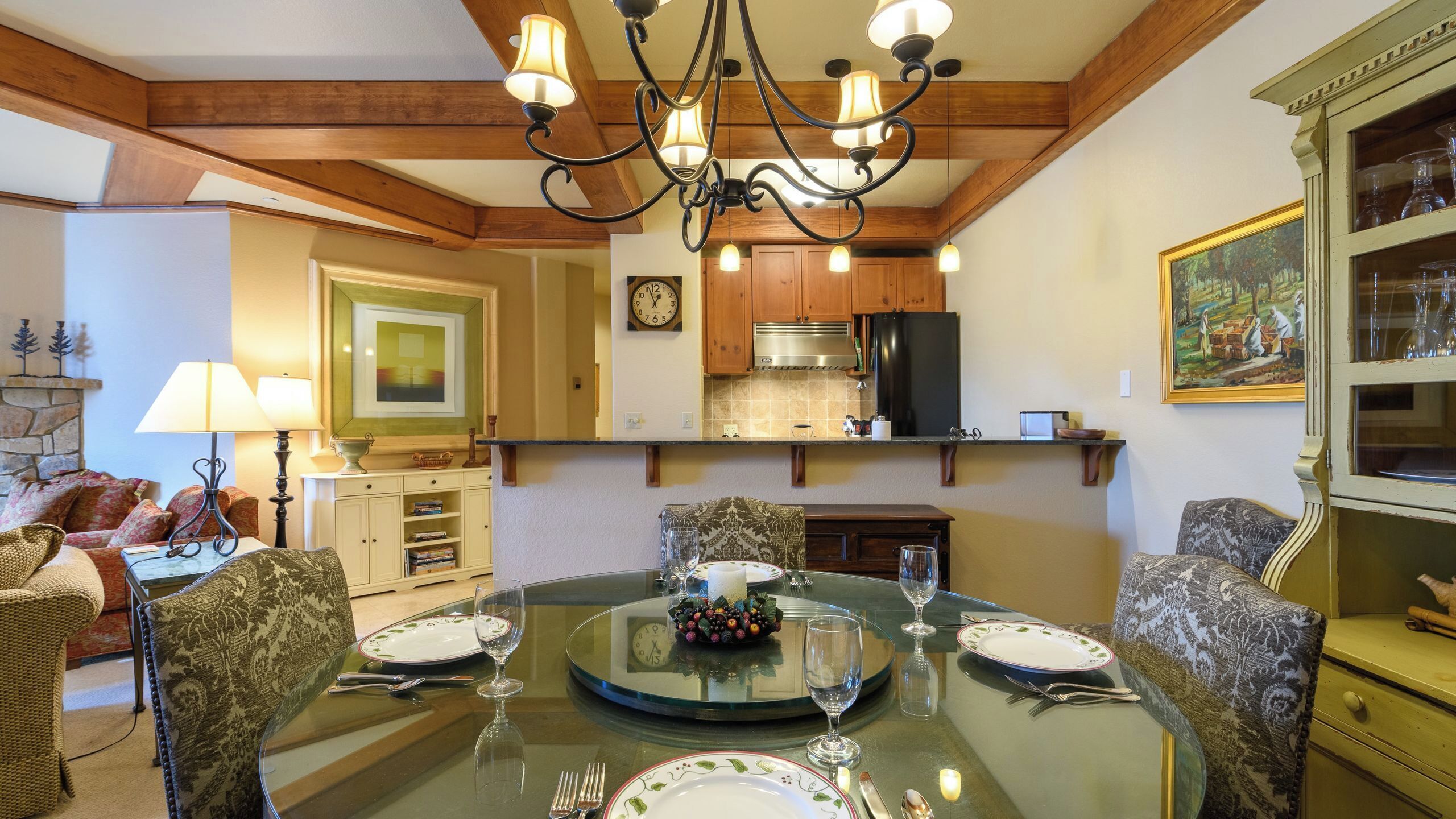 A dining area with a glass table set for four, surrounded by patterned chairs. A chandelier hangs above, and wooden beams and cabinets are visible in the background.