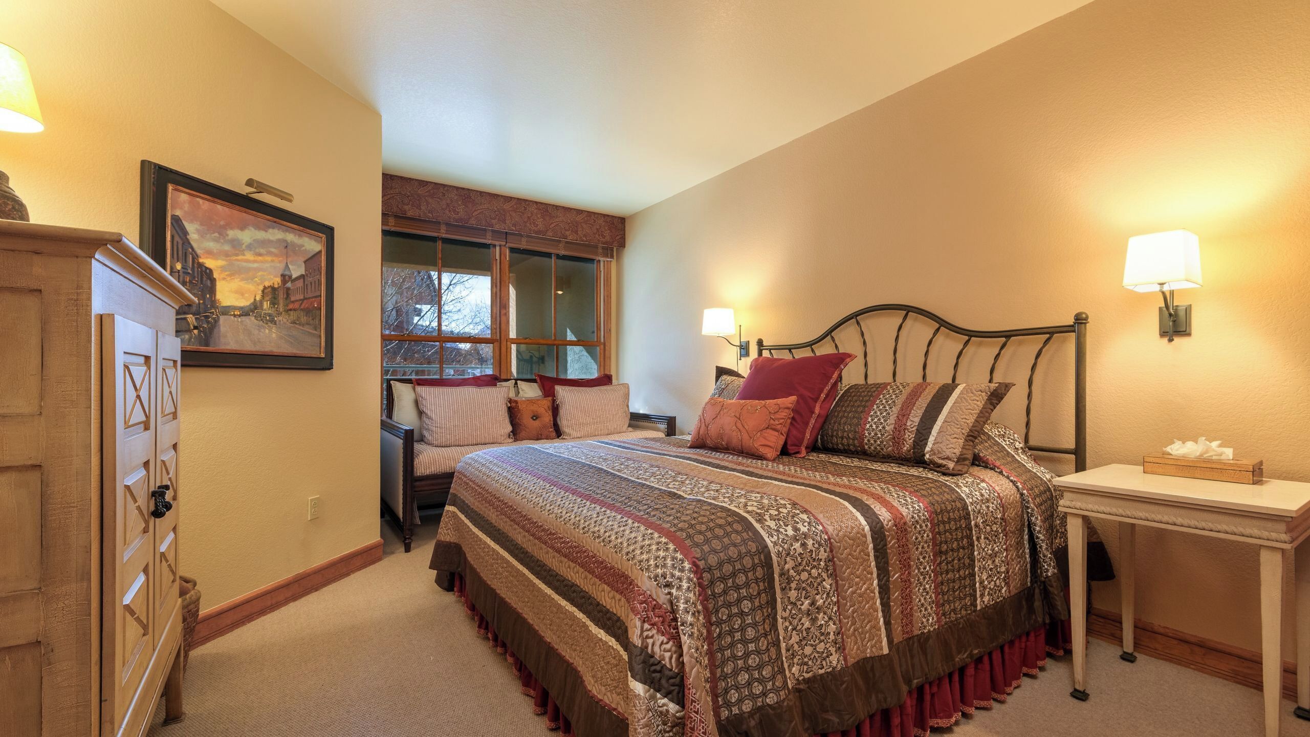 A bedroom with a large bed, a nightstand, a dresser, a window with a view, and a painting on the wall.