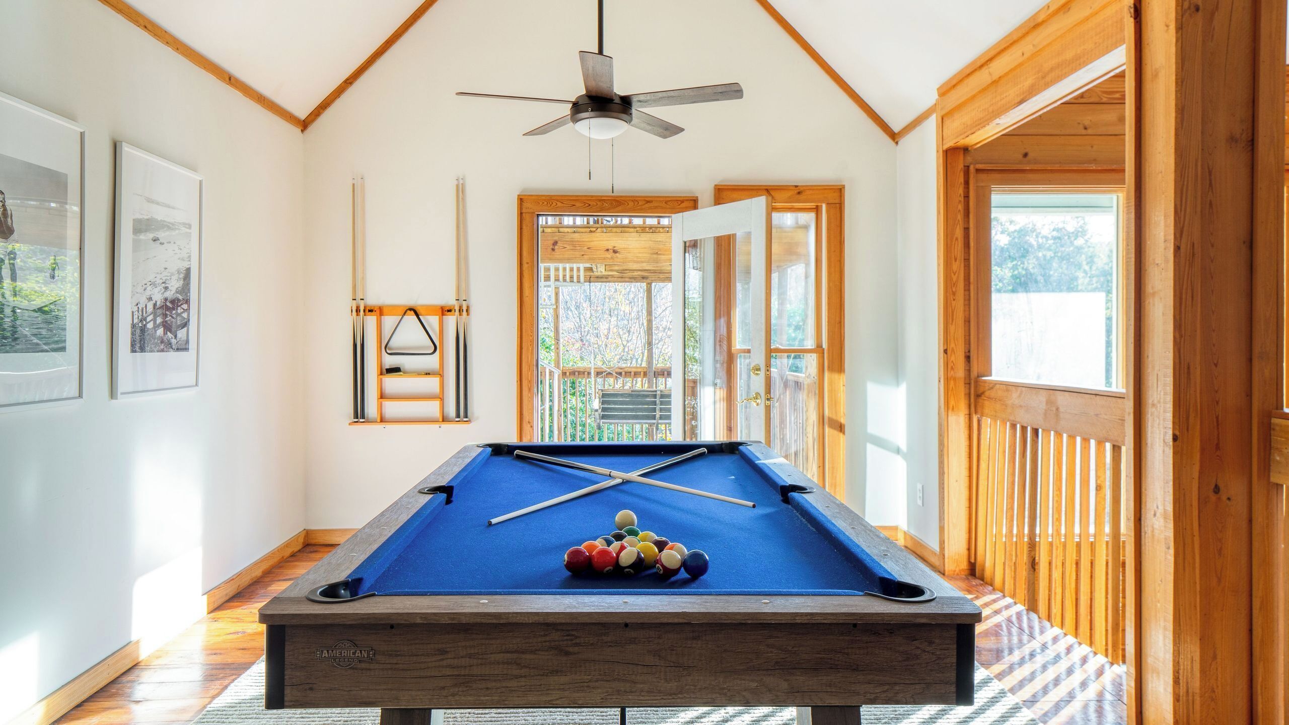 A pool table with a ceiling fan, framed pictures on the wall, and a wooden deck visible through the door.